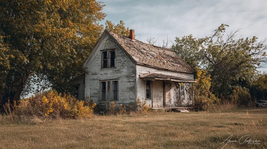 An old, weathered house with peeling paint and a collapsing porch, surrounded by overgrown grass and trees