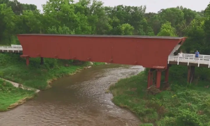 A red covered bridge spans a gentle stream, surrounded by lush green foliage