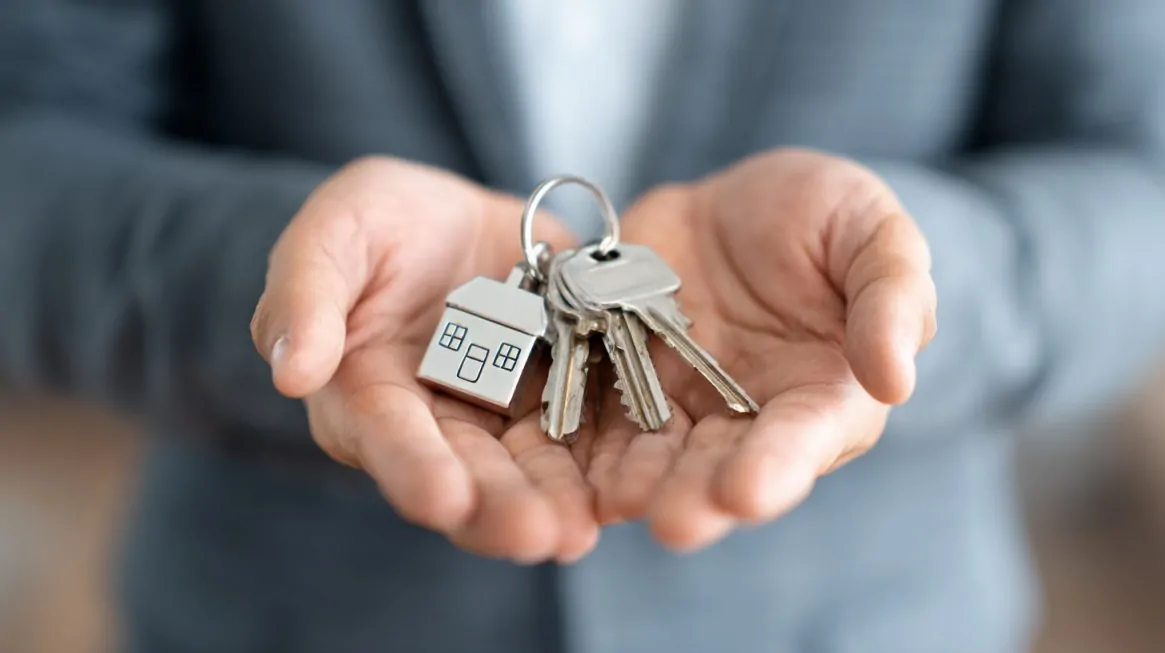 Close-up of hands offering house keys with a small house shaped keychain